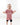 The image shows a young toddler walking indoors on a light-colored wooden floor. The child is smiling brightly and wearing a mauve-pink oversized T-shirt with black shorts underneath. The T-shirt has white lettering across the front. The toddler has a white bow in their hair and appears to be mid-step, looking joyful and energetic. The background is minimal and softly lit, creating a clean and airy atmosphere.