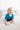 The image shows a young toddler with light curly hair crawling on a soft, neutral-colored bed. The child is looking slightly upward with a gentle smile, wearing a bright blue T-shirt and black pants. The setting appears to be a clean, minimal bedroom with a white wall and light bedding, creating a cozy and calm atmosphere. Natural light softly illuminates the scene.