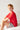 The image shows a young boy with red hair and freckles sitting on a soft surface against a light-colored wall. He is wearing a red short-sleeve shirt and light-colored shorts featuring a playful, multicolored pattern. He is smiling slightly and looking at the camera, with his arms resting on his knees in a relaxed pose. The setting appears bright and minimal.