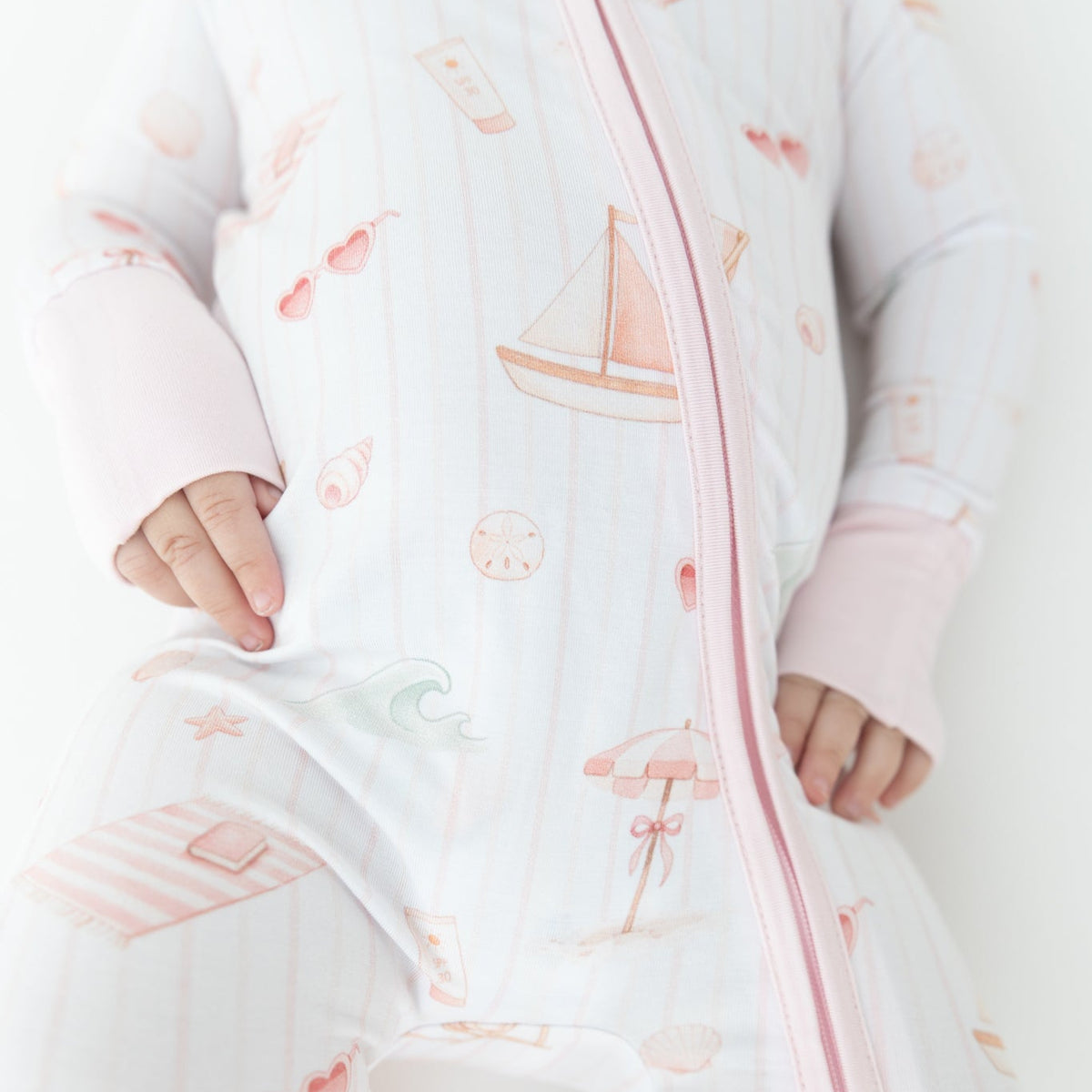 A baby wearing a white zip-up sleeper with pink trim and a beach-themed print featuring sailboats, umbrellas, and heart sunglasses.