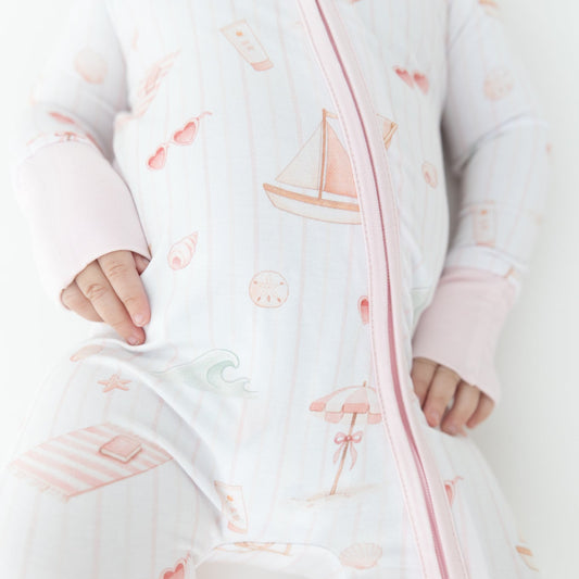 A baby wearing a white zip-up sleeper with pink trim and a beach-themed print featuring sailboats, umbrellas, and heart sunglasses.