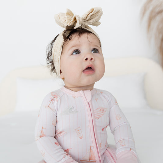 A baby wearing a white with light pink long-sleeve zip-up sleeper featuring sail boat, ocean waves and other beach onjects, looking up and to the side in a bright, clean setting.