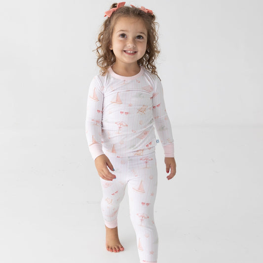A young girl with curly hair wearing a white long-sleeve pajamas with a pink and orange beach pattern, standing against a plain white studio background.