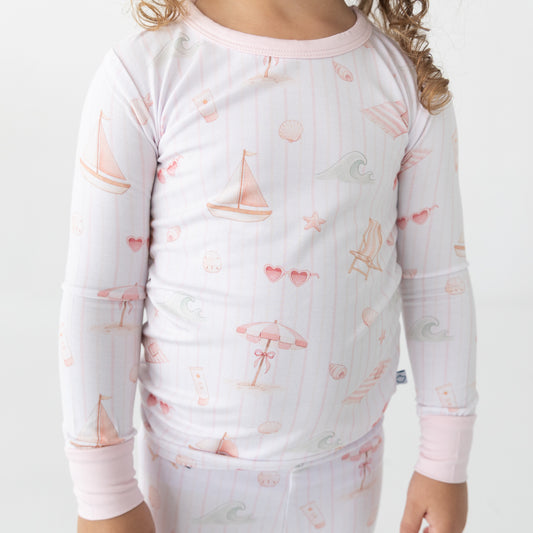A child wearing a long-sleeve pajamas with a pastel beach-themed print, including sailboats, umbrellas, hearts, and waves on a white background with thin pink vertical stripes.