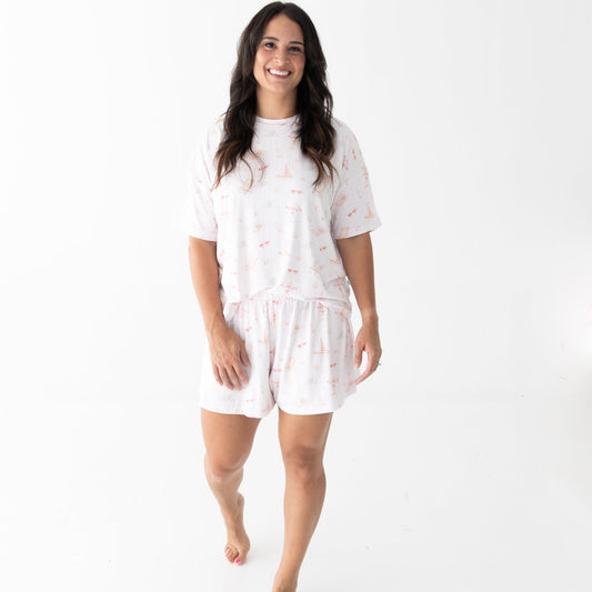 A woman modeling a matching white short-sleeve pajama top and shorts set with a subtle pink beach theme pattern, walking barefoot against a plain white studio background.
