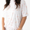 A woman wearing a white short-sleeve pajama top and matching shorts set with a delicate pink nautical print of sailboats and umbrellas.