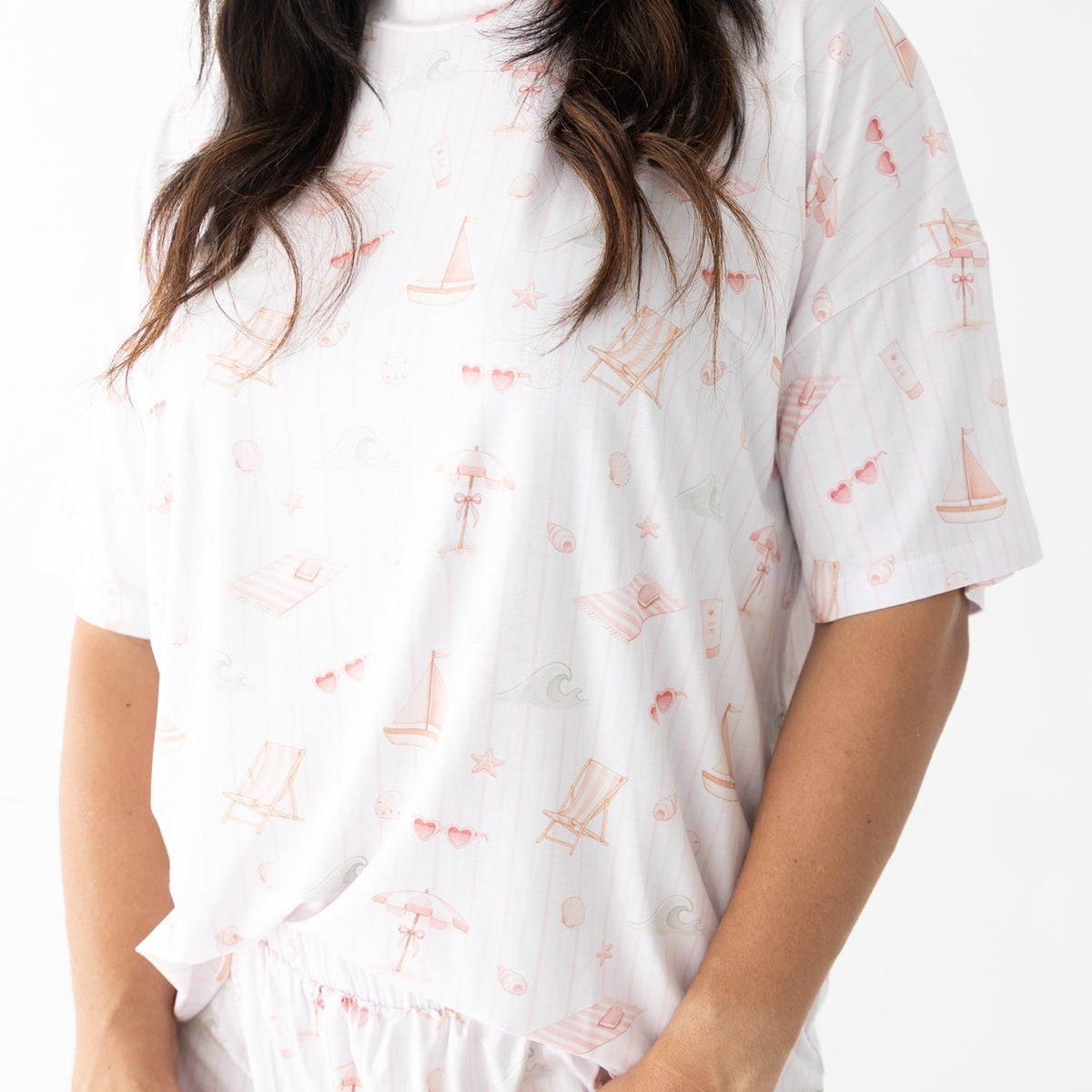 A woman wearing a white short-sleeve pajama top and matching shorts set with a delicate pink nautical print of sailboats and umbrellas.