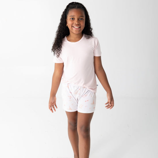 A young girl with curly  hair smiling and walking forward while wearing a light pink short-sleeve t-shirt and matching light pink shorts with a subtle beach pattern, set against a plain white studio background.