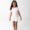 A young girl with curly  hair smiling and walking forward while wearing a light pink short-sleeve t-shirt and matching light pink shorts with a subtle beach pattern, set against a plain white studio background.