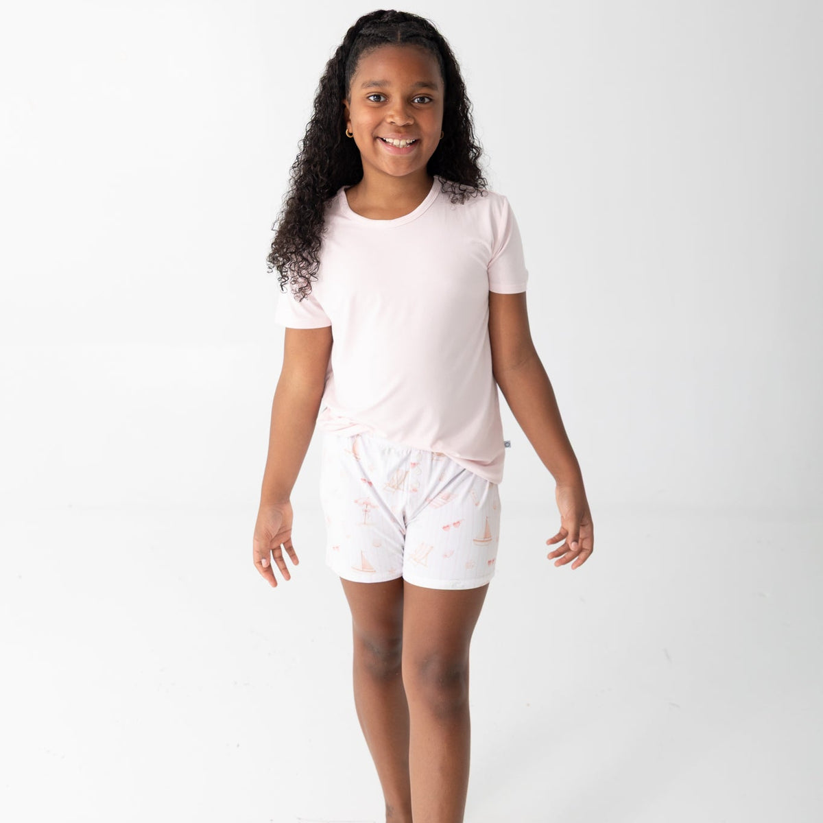 A young girl with curly  hair smiling and walking forward while wearing a light pink short-sleeve t-shirt and matching light pink shorts with a subtle beach pattern, set against a plain white studio background.
