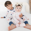 Two children sitting on a white bed wearing beach patterned pajamas. The older child is a boy with brown hair wearing a navy and white pajama set with small red and orange ocean accents, and the younger child is a girl wearing a light pink pajama set with sail boat, umbrella beach objects.