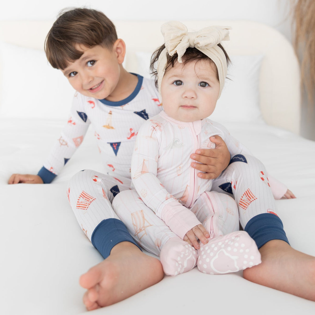 Two children sitting on a white bed wearing beach patterned pajamas. The older child is a boy with brown hair wearing a navy and white pajama set with small red and orange ocean accents, and the younger child is a girl wearing a light pink pajama set with sail boat, umbrella beach objects.