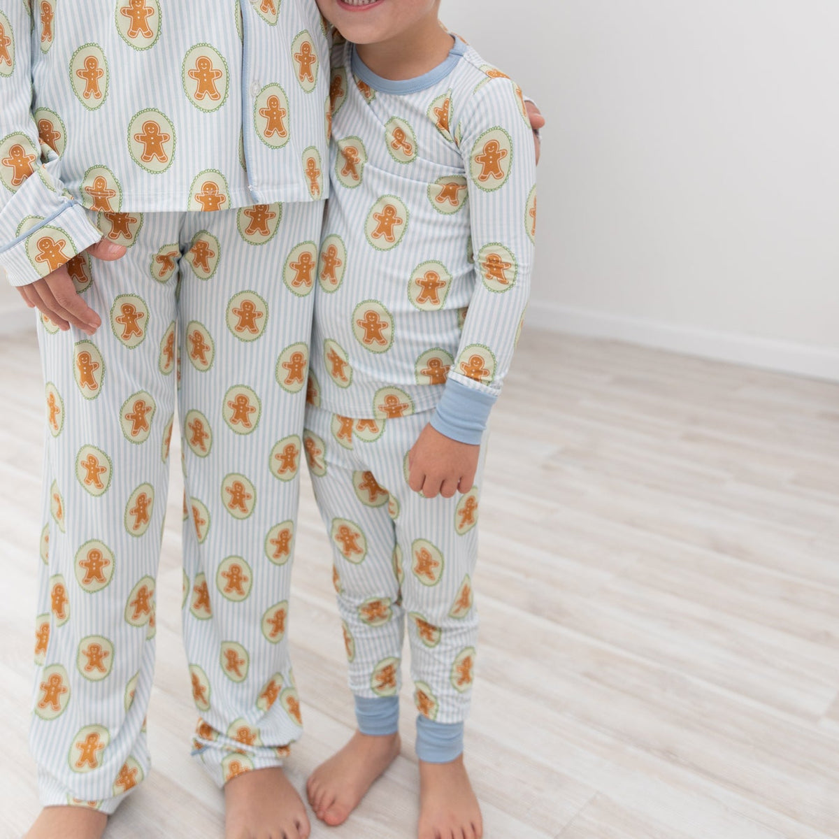 A young child wearing a light blue and white striped pajama set with an orange gingerbread man pattern, standing next to an adult wearing a matching pajama top and pants in a bright room with light wood floors.