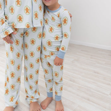 A young child wearing a light blue and white striped pajama set with an orange gingerbread man pattern, standing next to an adult wearing a matching pajama top and pants in a bright room with light wood floors.