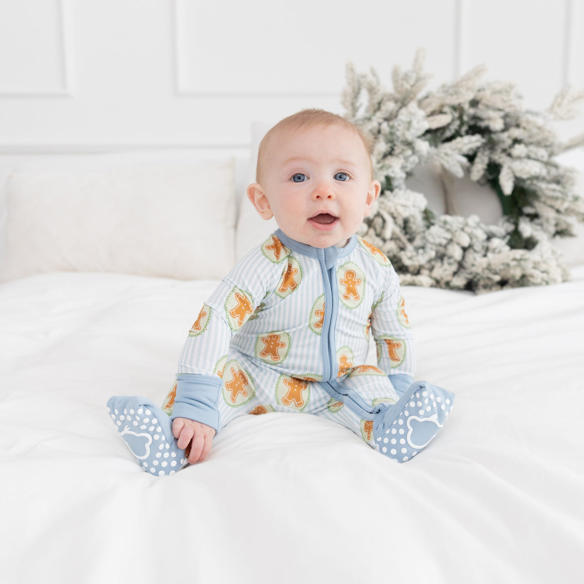 A baby sitting on a white bed wearing a light blue and white striped footless sleeper with gingerbread patterns, featuring white polka-dot monkey grippers.