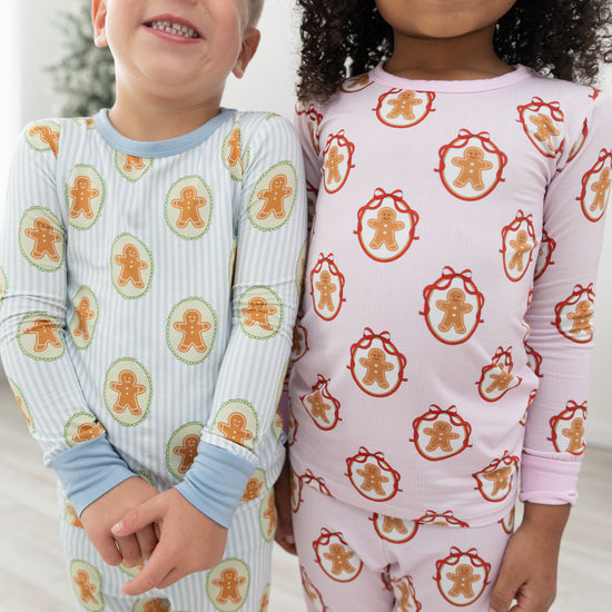 Two children wearing long-sleeve pajamas with gingerbread-themed prints. The child on the left wears light blue and white striped pajamas with orange gingerbread cookies, and the child on the right wears a pale pink pajama top and matching bottoms with red gingerbread cookie motifs.