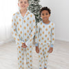 Two children standing barefoot in a bright room wearing matching long-sleeve pajamas with a light blue and white striped base and a repeating gingerbread man pattern. A decorated Christmas tree is visible in the background.