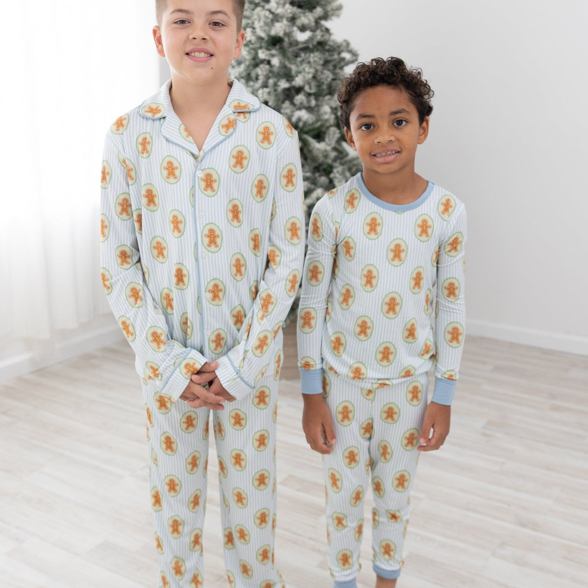 Two children standing barefoot in a bright room wearing matching long-sleeve pajamas with a light blue and white striped base and a repeating gingerbread man pattern. A decorated Christmas tree is visible in the background.