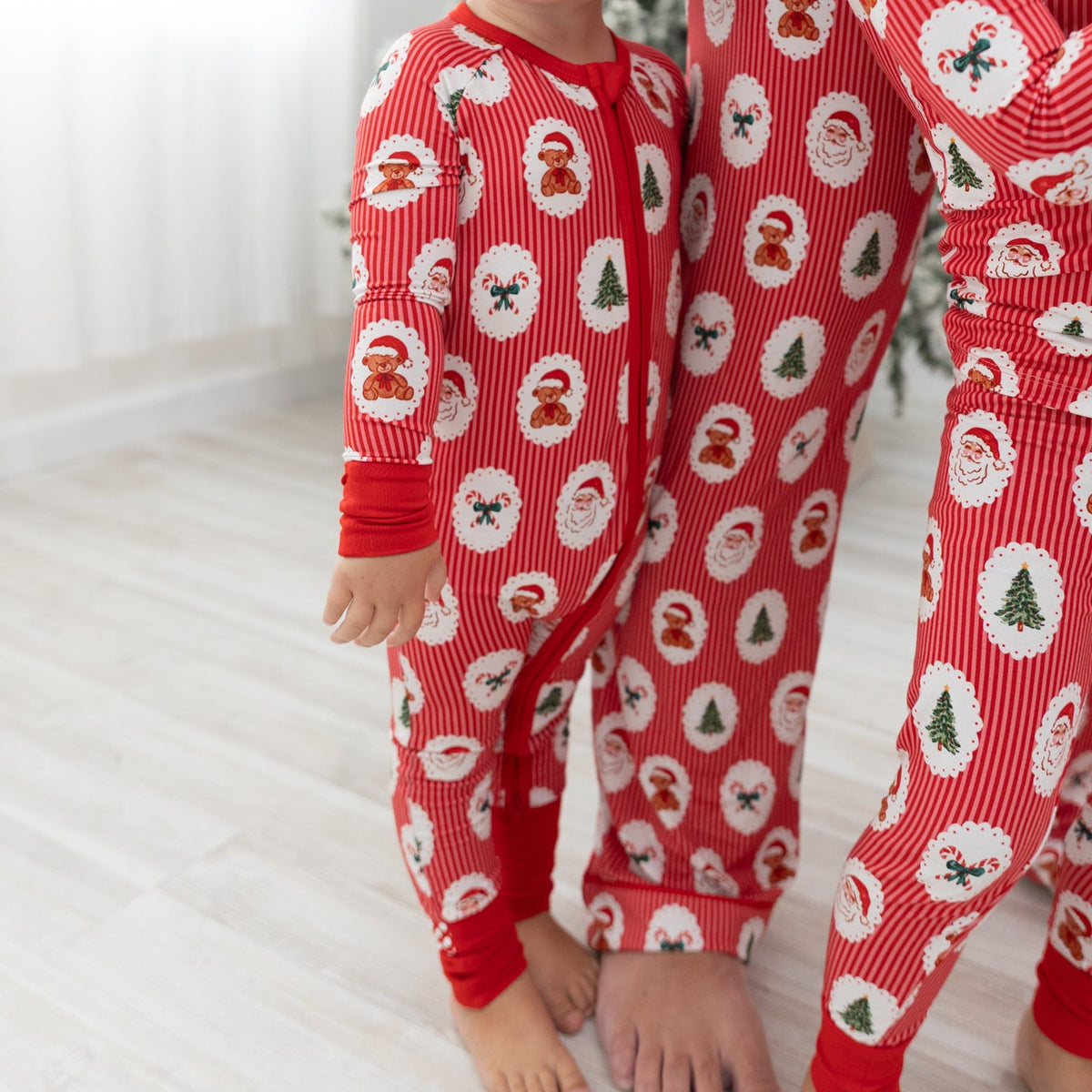 A toddler wearing a matching red Christmas pajamas with a Santa Claus, christmas tree, candy cane, teddy bear repeating pattern, standing barefoot on a light wood floor next to an adult wearing matching pajamas.