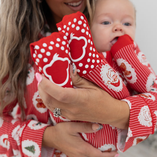 A close-up lifestyle shot of a baby wearing red and white striped Christmas pajamas wearing featuring a Santa Claus, christmas tree, candy cane, teddy bear repeating pattern. Infant feet feature a white monkey logo with polka dot grippers, held by a woman wearing matching red and white patterned long-sleeve pajamas.