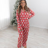 A woman stands barefoot in a bright room wearing red Christmas pajamas set with a Santa Claus, christmas tree, candy cane, teddy bear repeating pattern. A flocked Christmas tree is visible in the background.