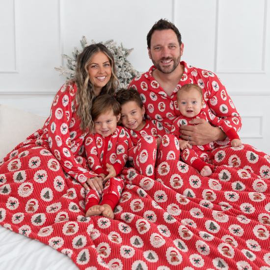 A family of four wearing matching red Christmas pajamas featuring a festive Christmas print with Santa Claus faces, teddy bears, candy canes, and trees on red-and-white stripes sitting together on a bed under a matching red blanket. 
