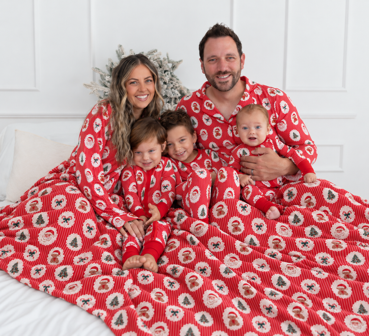 A family of four wearing matching red Christmas pajamas featuring a festive Christmas print with Santa Claus faces, teddy bears, candy canes, and trees on red-and-white stripes sitting together on a bed under a matching red blanket. 