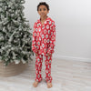 A young boy wearing a red long-sleeve pajama set with christmas trees, candy canes, Santa Claus pattern, standing barefoot in a room next to a flocked Christmas tree.