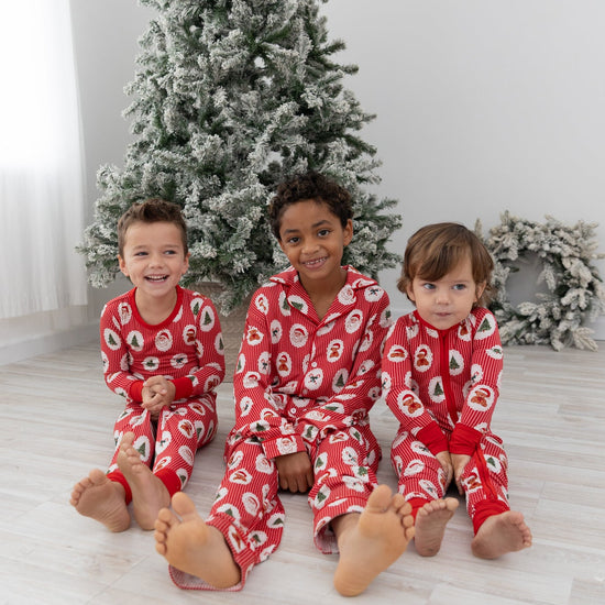 Three children sitting barefoot on a light wood floor in front of a decorated Christmas tree, wearing matching red holiday pajamas with a santa clause, christmas trees and candy cane pattern