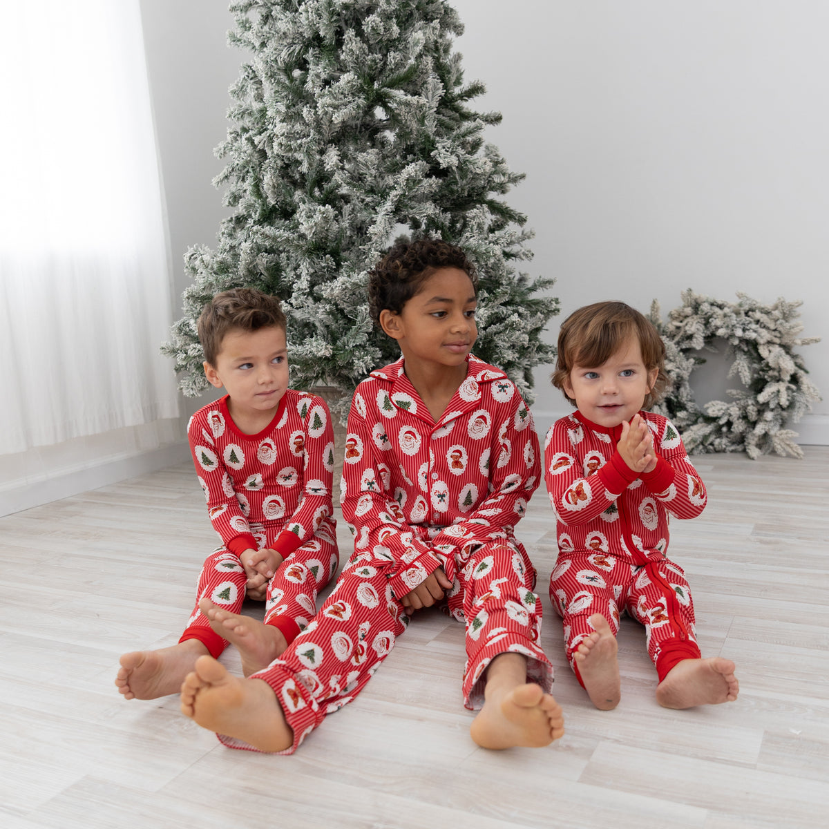 Three children sitting on a light wood floor in front of a decorated Christmas tree, wearing matching red holiday pajamas with a Santa Claus, christmas tree, candy cane, teddy bear repeating pattern.