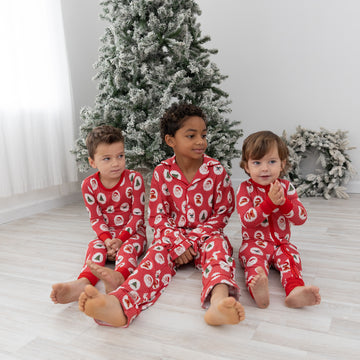 Three children sitting on a light wood floor in front of a decorated Christmas tree, wearing matching red holiday pajamas with a Santa Claus, christmas tree, candy cane, teddy bear repeating pattern.