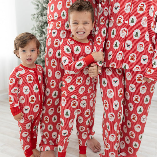 A family of four wearing matching red Christmas pajamas with a Santa Claus, christmas tree, candy cane, teddy bear repeating pattern, standing barefoot in a brightly lit room with a Christmas tree in the background.