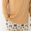 A person wearing a mustard yellow long-sleeve top and matching pants with a Halloween-themed print. The pajamas feature a cream base with vertical stripes and illustrations of ghosts, pumpkins, a haunted house, bats, spiders, and stars.