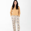 A Woman wearing a mustard yellow long-sleeve top and matching pants with a Halloween-themed print. The pajamas feature a cream base with vertical stripes and illustrations of ghosts, pumpkins, a haunted house, bats, spiders, and stars.