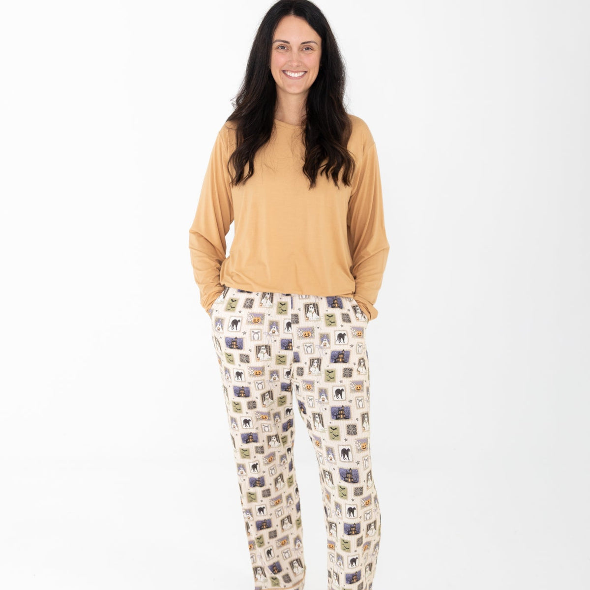 A Woman wearing a mustard yellow long-sleeve top and matching pants with a Halloween-themed print. The pajamas feature a cream base with vertical stripes and illustrations of ghosts, pumpkins, a haunted house, bats, spiders, and stars.