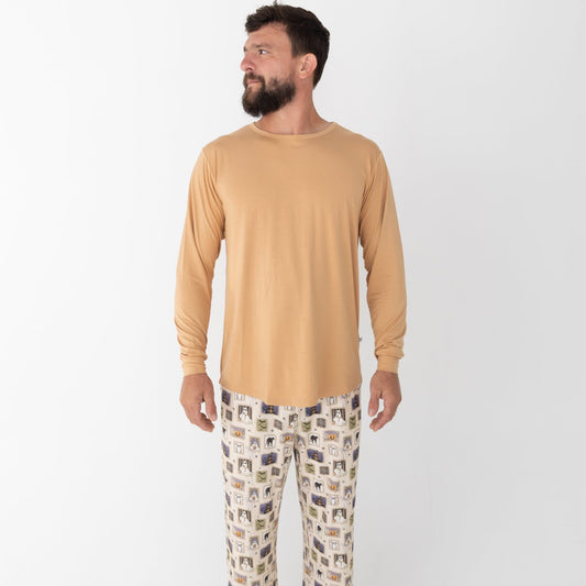 A man wearing a mustard yellow long-sleeve top and matching pants with a Halloween-themed print. The pajamas feature a cream base with vertical stripes and illustrations of ghosts, pumpkins, a haunted house, bats, spiders, and stars.