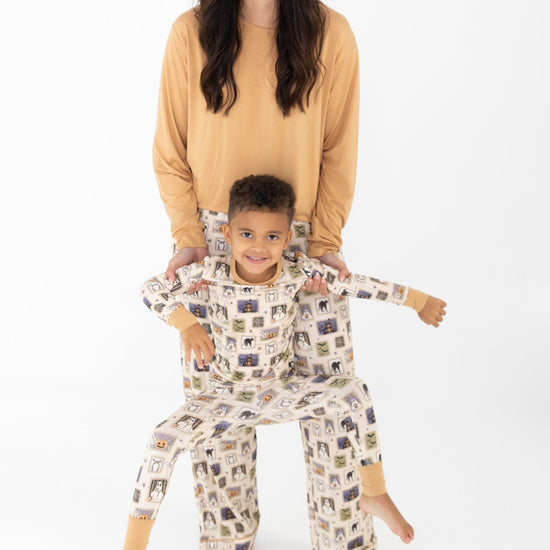 A woman and a young boy wearing matching pajamas, with the woman standing behind the boy and holding his arms. The boy's pajamas feature a long-sleeve pajama top and matching pants with a Halloween-themed print. The pajamas feature a cream base with vertical stripes and illustrations of ghosts, pumpkins, a haunted house, bats, spiders, and stars. woman wears a solid mustard yellow long-sleeve top and long pants that match the boys pajama prints.