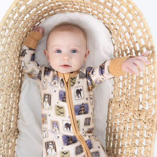 A baby wearing a beige base halloween-print featuring stamps of ghosts, black cat, bows, bats, spider webs, jack-o-lantern zippy romper, lies inside a woven rattan bassinet on a white background.