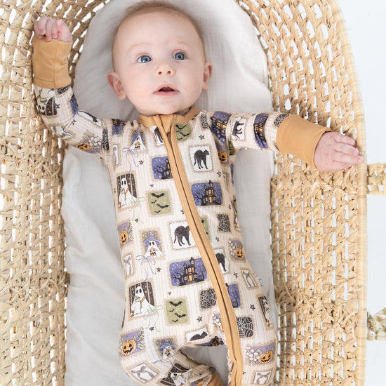 A baby lying on their back inside a woven rattan bassinet, wearing a beige base halloween-print featuring stamps of ghosts, black cat, bows, bats, spider webs, jack-o-lantern