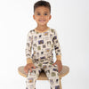 A young boy sitting on a wooden stool wearing a beige base halloween-print featuring stamps of ghosts, black cat, bows, bats, spider webs, jack-o-lantern.