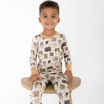 A young boy sitting on a wooden stool wearing a beige base halloween-print featuring stamps of ghosts, black cat, bows, bats, spider webs, jack-o-lantern.