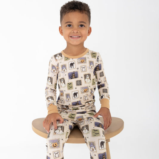 A young boy sitting on a wooden stool wearing a beige base halloween-print featuring stamps of ghosts, black cat, bows, bats, spider webs, jack-o-lantern.