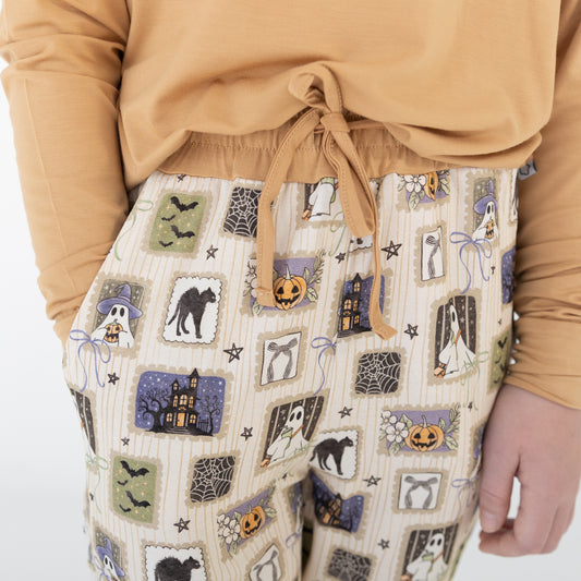 A person wearing a mustard yellow long-sleeve top and matching pants with a Halloween-themed print. The pajamas feature a cream base with vertical stripes and illustrations of ghosts, pumpkins, a haunted house, bats, spiders, and stars.