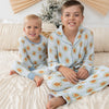 Two children sitting on a bed wearing matching light blue pajamas with a repeating gingerbread cookie print. The room has a warm neutral color palette with a beige wall, a decorative fan, and a small frosted tree in the background.