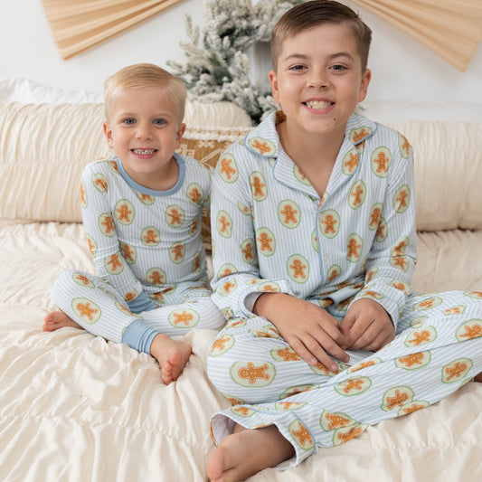 Two children sitting on a bed wearing matching light blue pajamas with a repeating gingerbread cookie print. The room has a warm neutral color palette with a beige wall, a decorative fan, and a small frosted tree in the background.