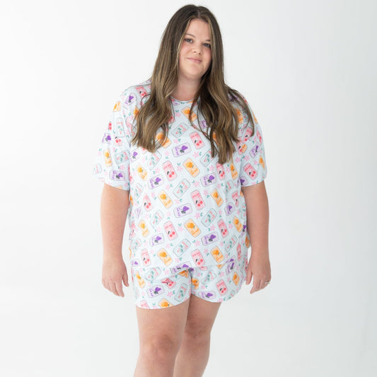 A woman modeling a matching short-sleeve pajama set with a colorful soda cans pattern on a light blue background, photographed against a seamless white studio backdrop.