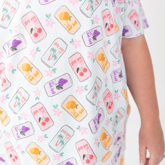 A Woman wearing a short-sleeve pajama top with a colorful repeating pattern of fruit and flowers, colorful soda cans pattern on a light blue background, photographed against a seamless white studio backdrop.