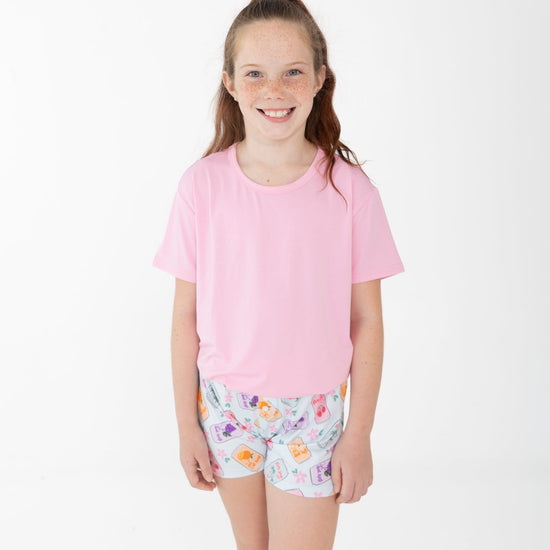 A young girl with her hair in a ponytail stands against a plain white studio background, wearing a light pink short-sleeve t-shirt and light blue shorts with a colorful floral and soda can pattern.