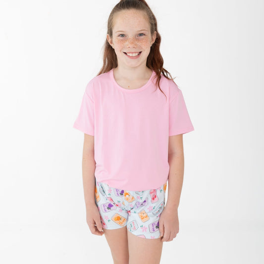 A young girl with her hair in a ponytail stands against a plain white studio background, wearing a light pink short-sleeve t-shirt and light blue shorts with a colorful floral and soda can pattern.