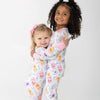 Two young children hugging while wearing matching long-sleeve pajamas with a light blue background with a colorful cherry, orange and grape soda cans print. set against a plain white studio background.